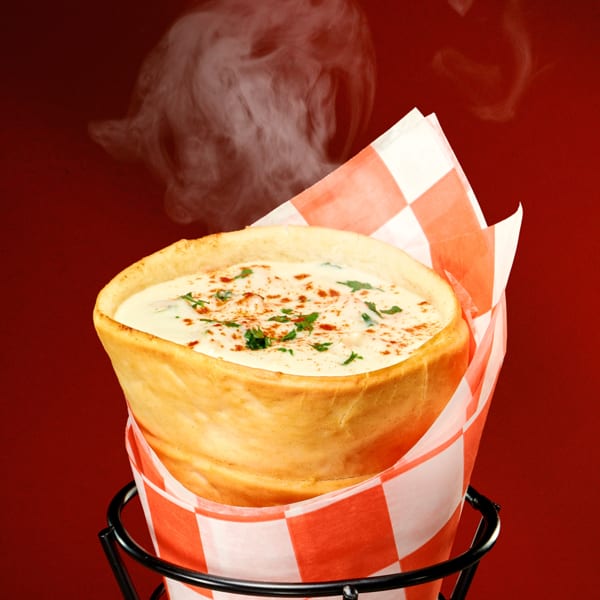 Steaming hot bread cone filled with creamy chowder, garnished with herbs and paprika, in a metal holder.