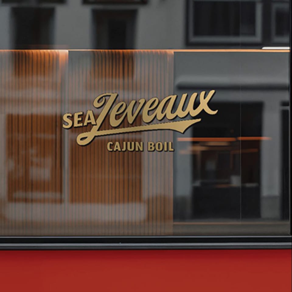 Golden "Sea Jeveaux Cajun Boil" logo on a frosted glass window with a red base.
