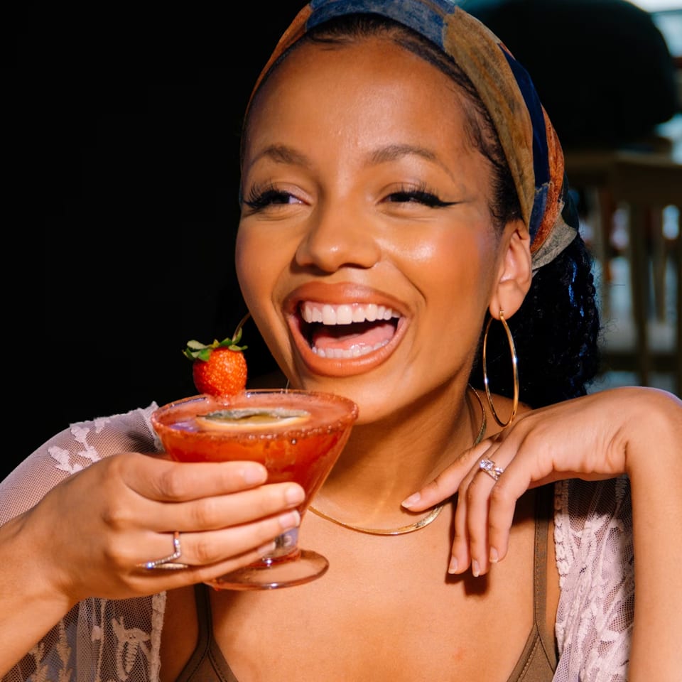 A smiling woman in a headscarf holds a strawberry cocktail, wearing hoop earrings and rings.